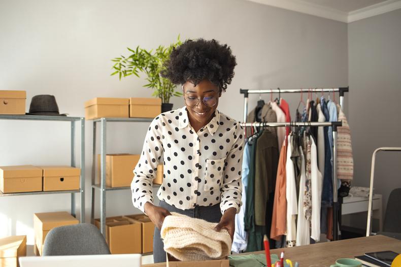 a woman packs clothing into a box on a table in front of a clothes rail and sheleves of boxes